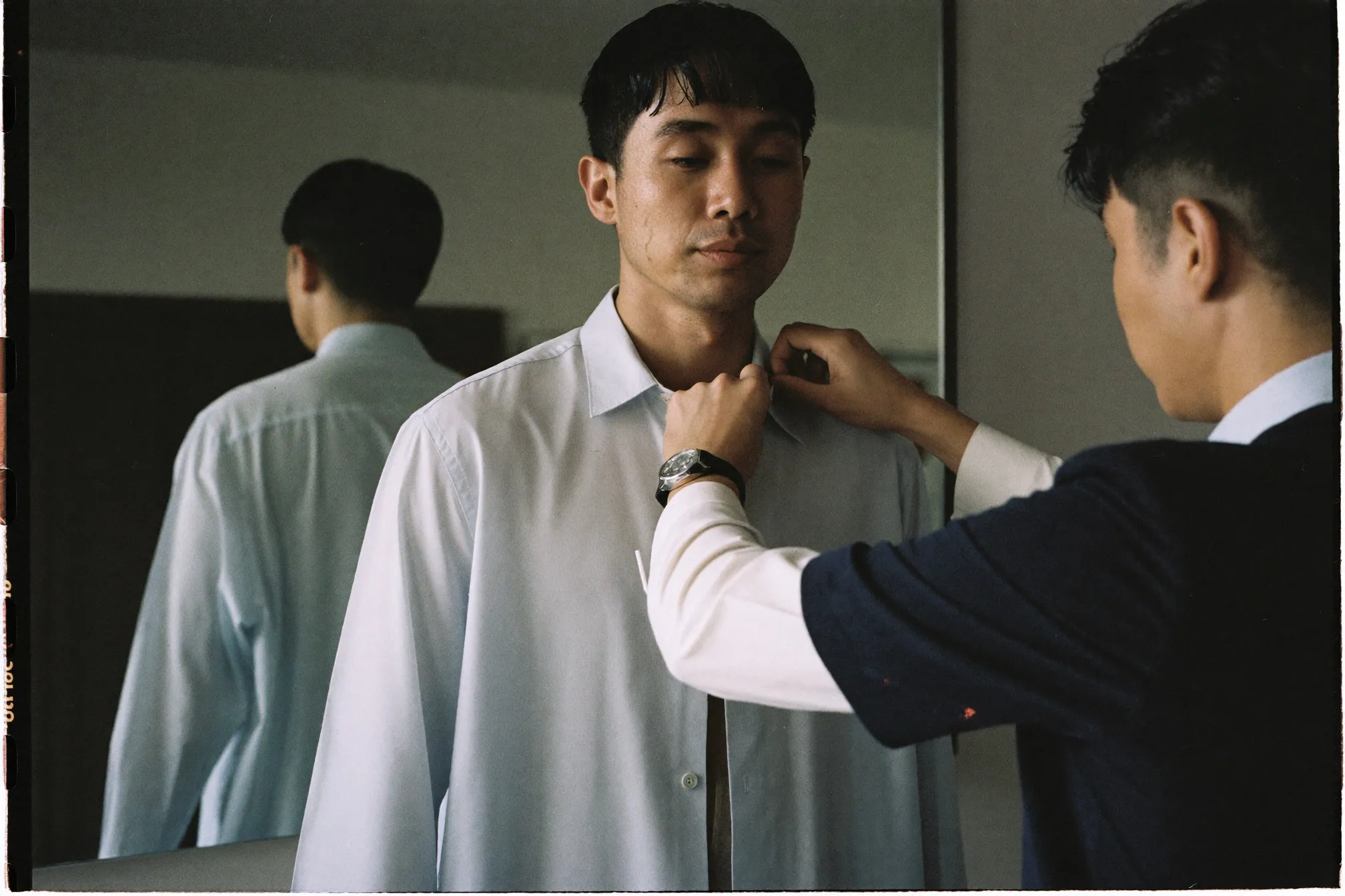 Man standing at a mirror during a shirt fitting while the tailor pins the side seam