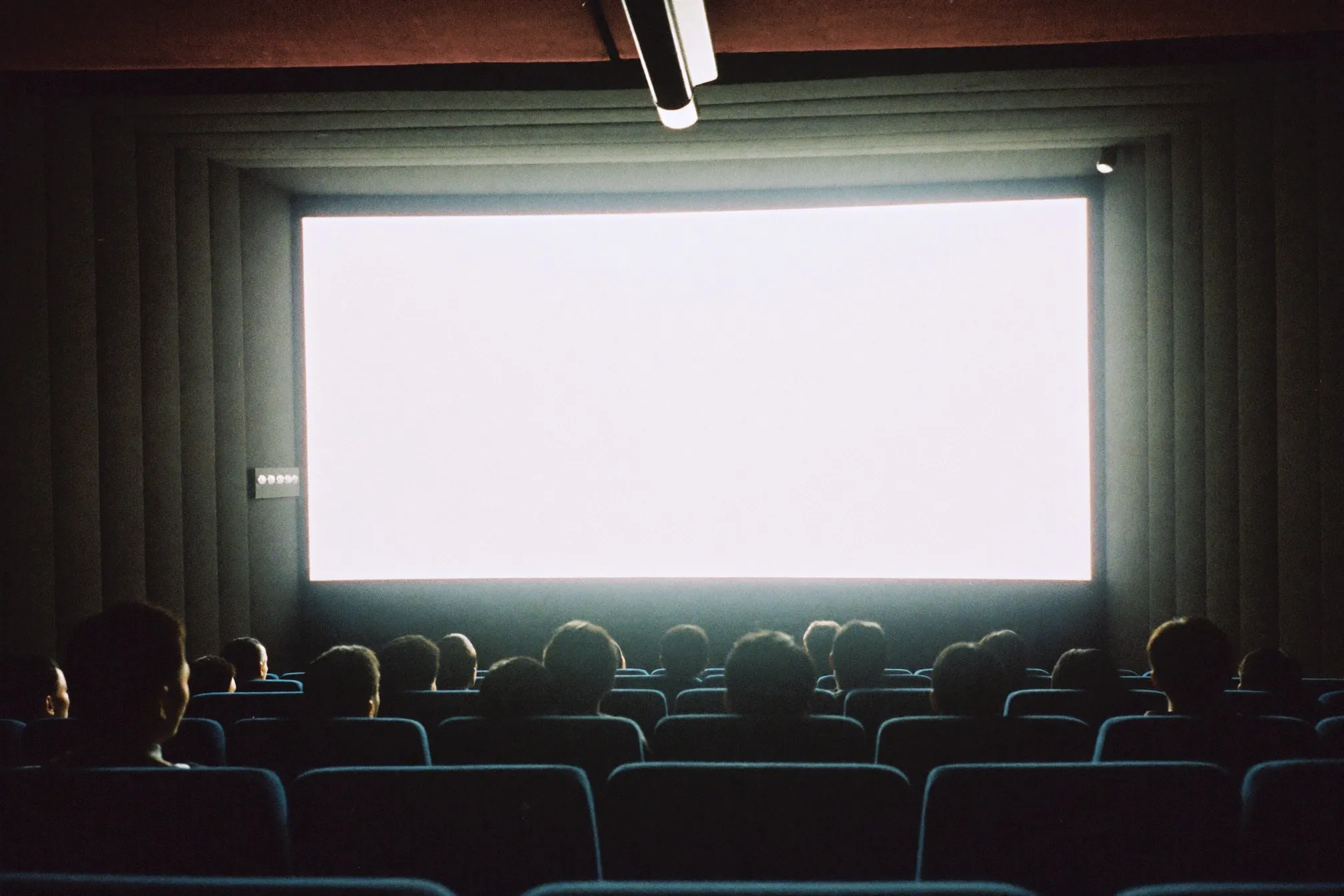 Packed cinema auditorium shot from the back row, audience silhouettes against a bright screen