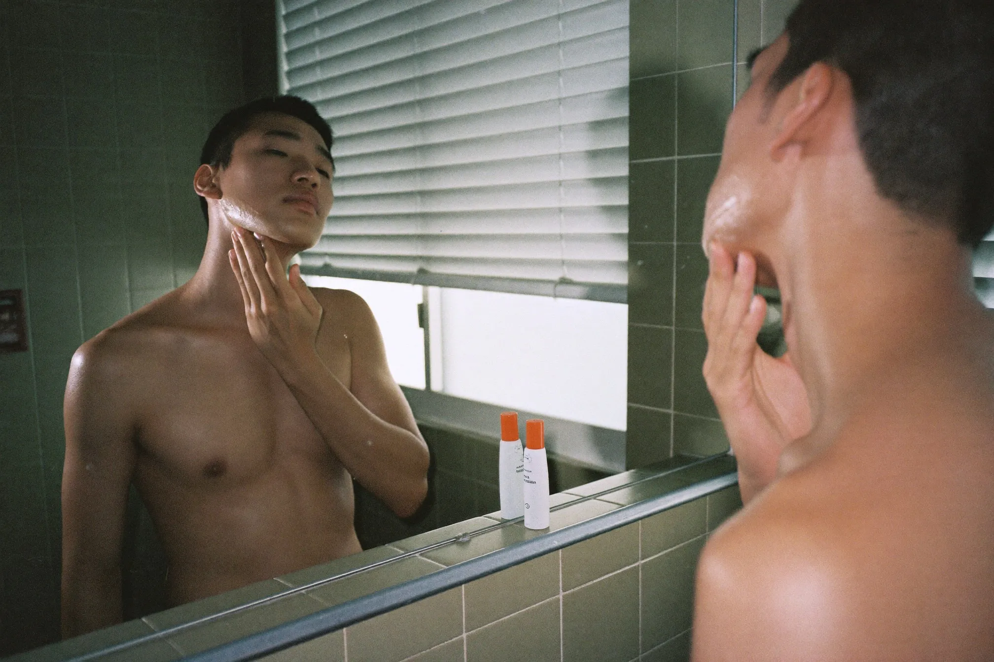 Man pressing tinted sunscreen along his jawline at a bathroom mirror in morning light