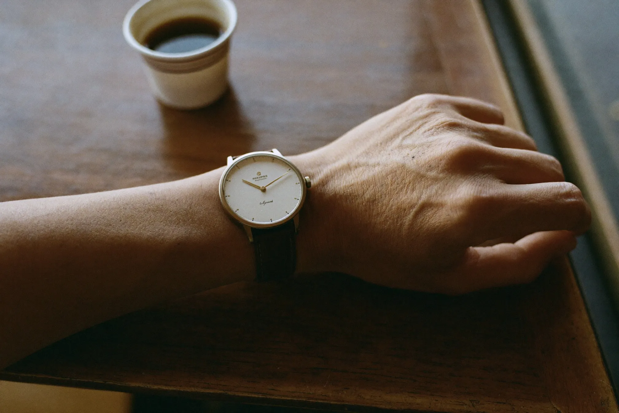 Close view of a generic unbranded automatic watch with a plain dial on a man's wrist beside a cup of kopi