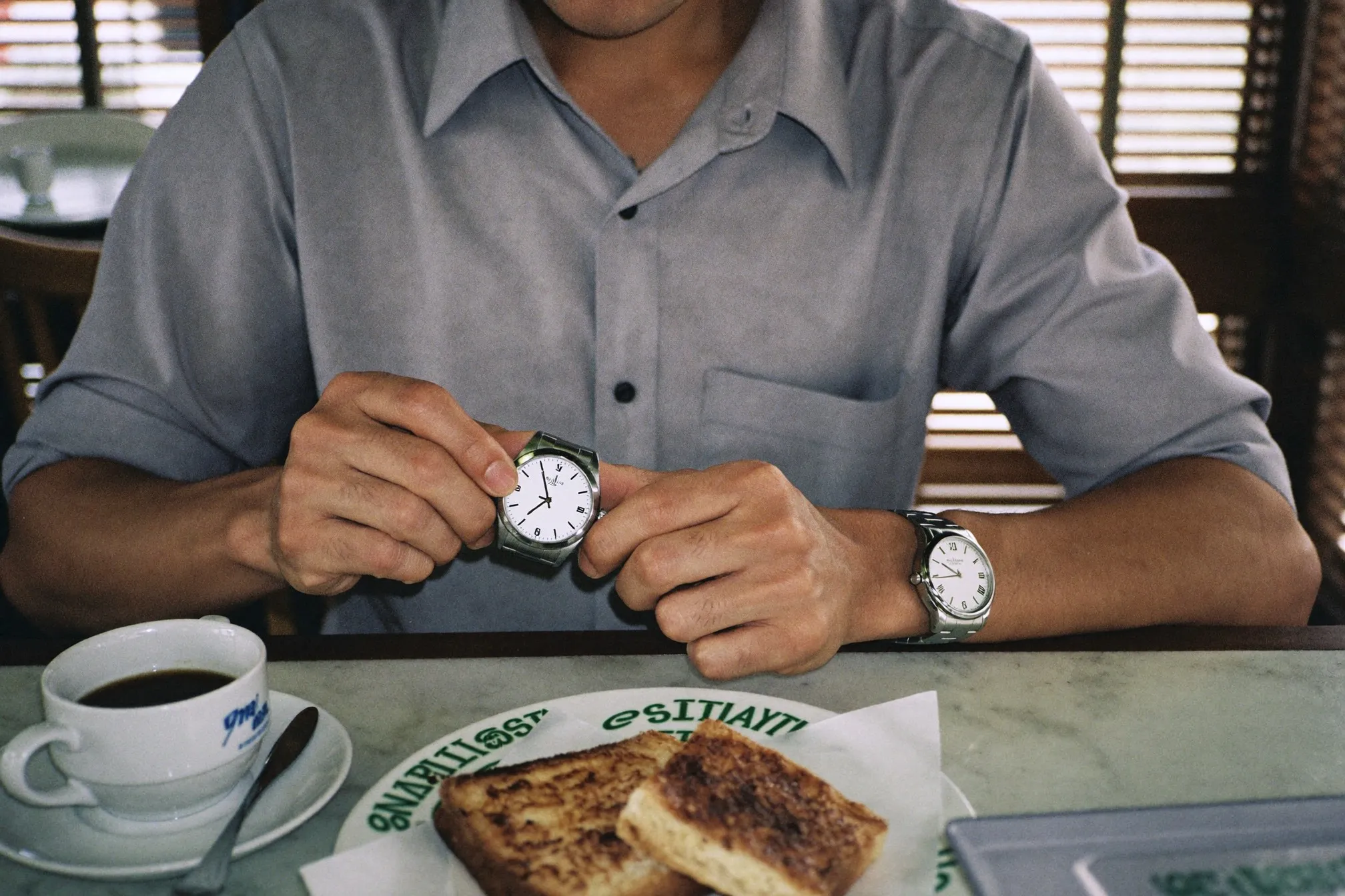 Man in a linen shirt checking a plain steel watch on his wrist at a kopitiam table in Kuala Lumpur
