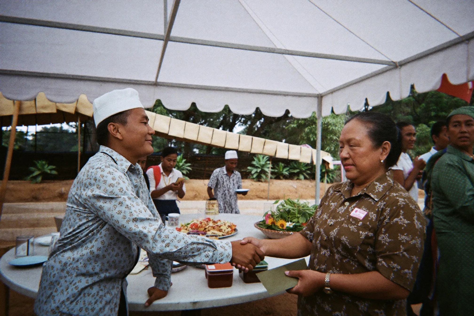 Guest in a batik shirt greeting the hosts and handing over a green envelope at an outdoor Malay wedding kenduri