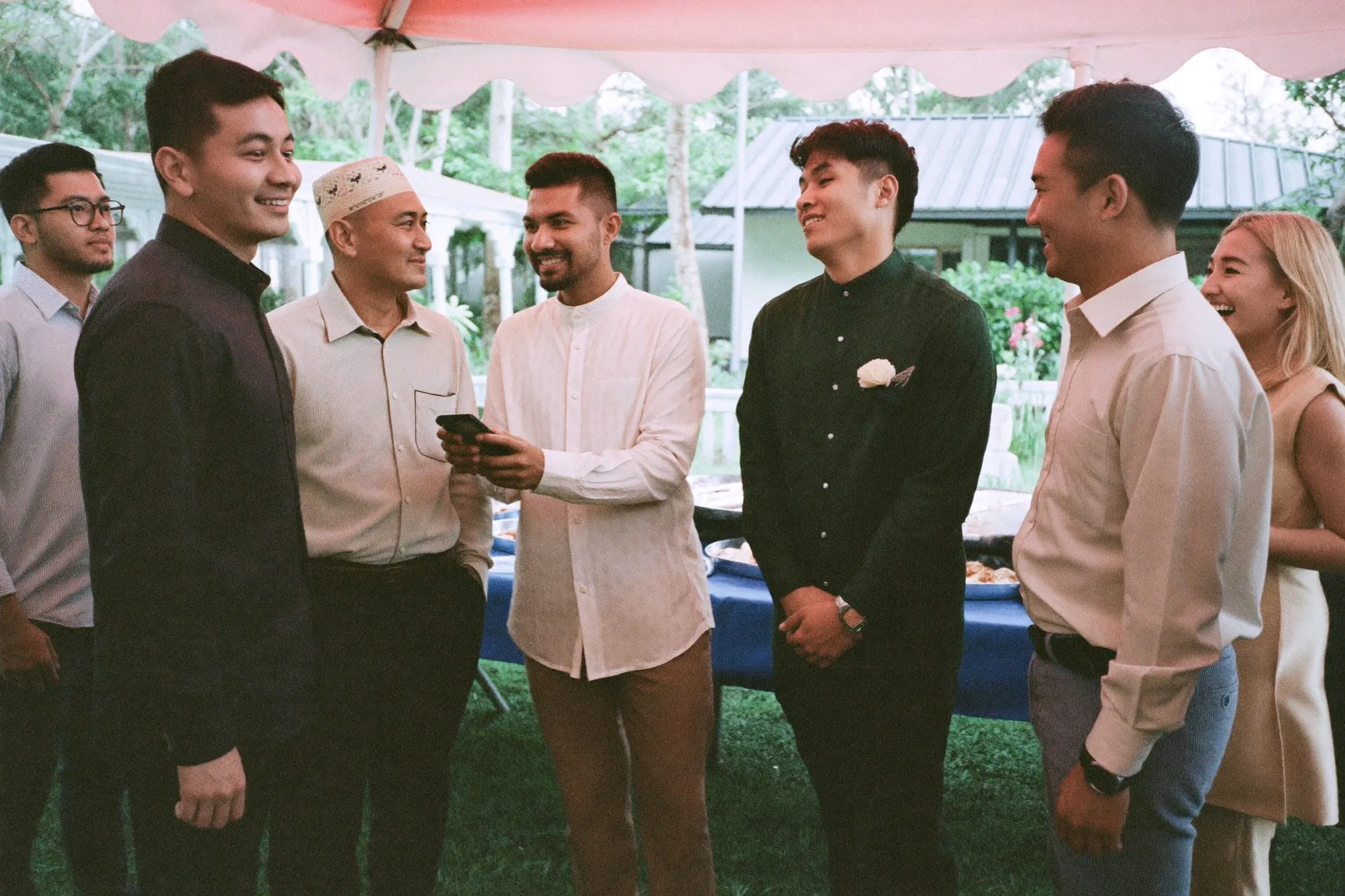 Wedding guests in long-sleeve shirts and a baju melayu chatting under a canopy at an outdoor kenduri in Malaysia