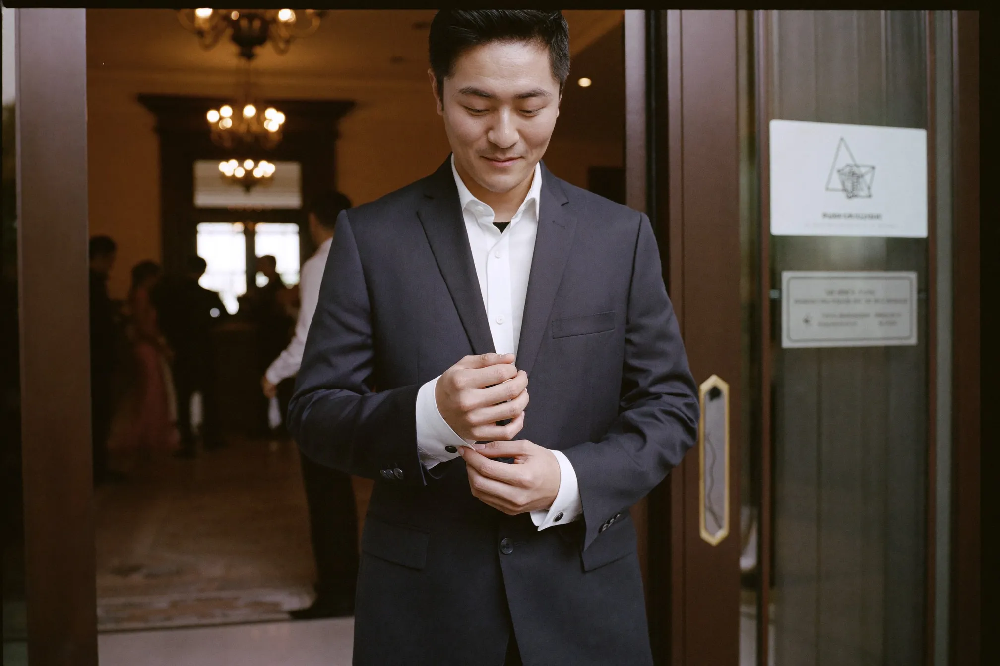Man in a navy unstructured blazer and white shirt adjusting his cuff outside a hotel ballroom wedding reception in Kuala Lumpur