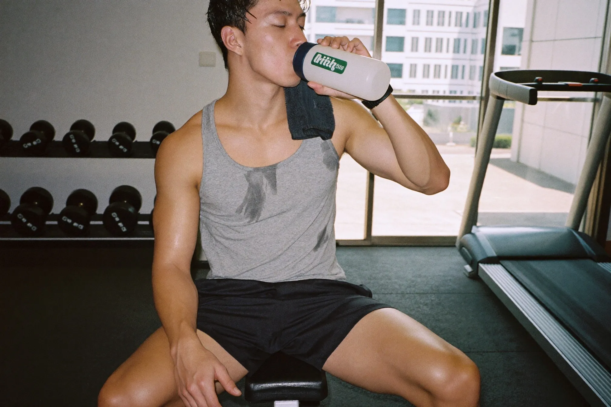 Malay man sitting on a bench in a condo gym drinking from a plain shaker bottle after training