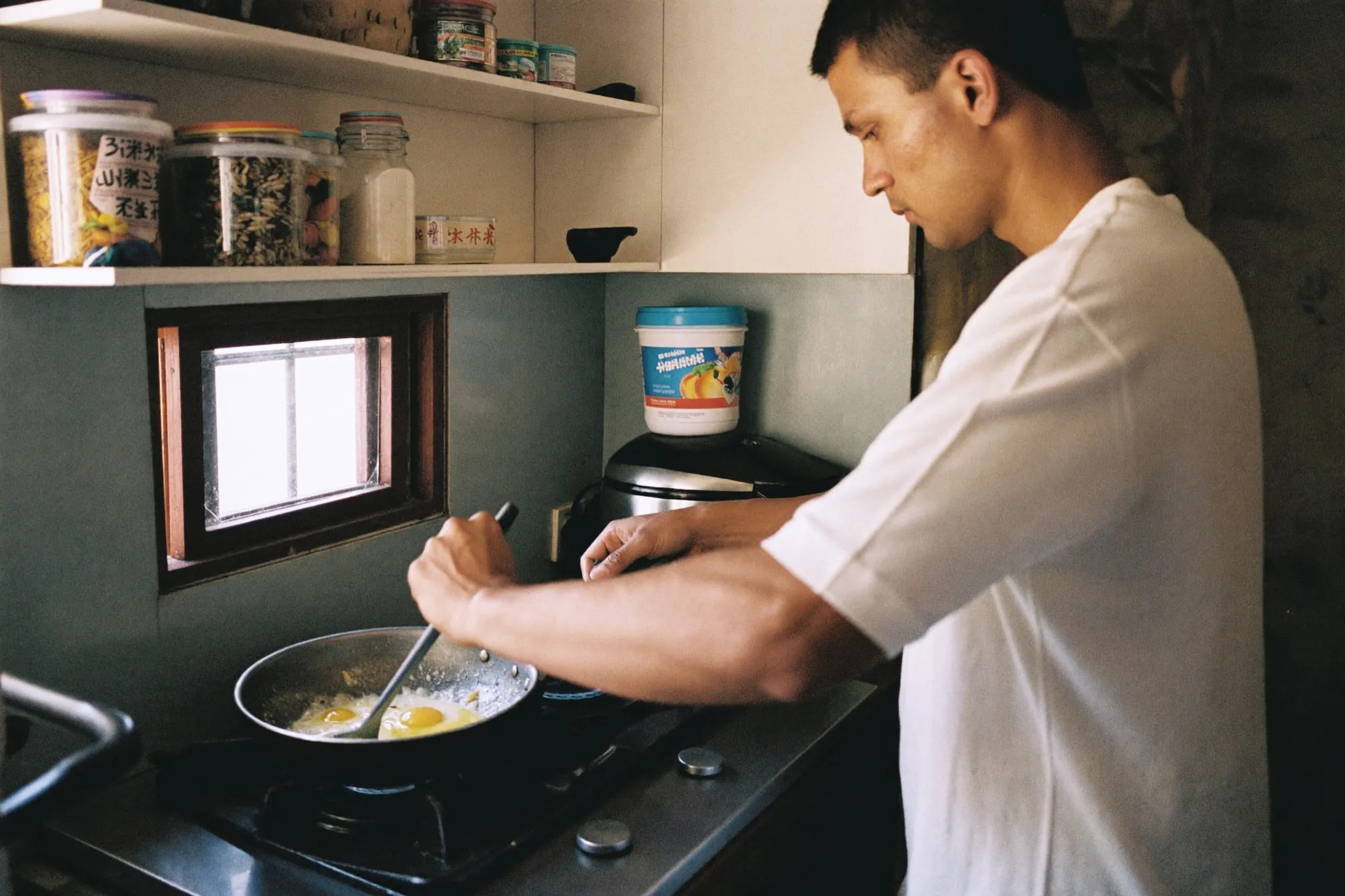 Indian Malaysian man cooking eggs in a small HDB kitchen with a plain protein tub on the counter behind him