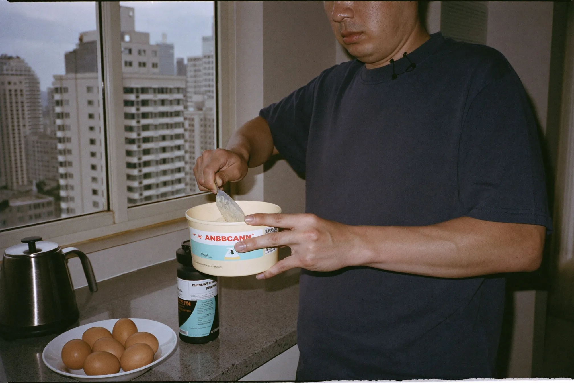 Chinese Malaysian man scooping protein powder into a plain shaker bottle on a kitchen counter in a KL apartment