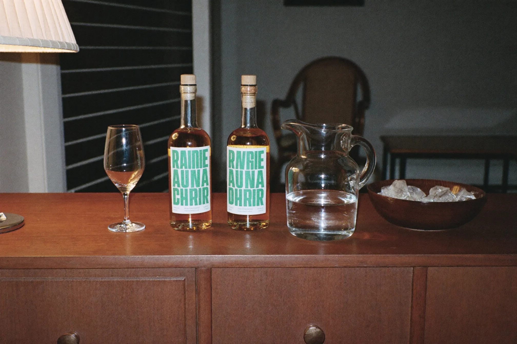 Two plain unlabelled bottles of amber spirit, a small water jug and a tulip glass on a sideboard in a Singapore HDB living room