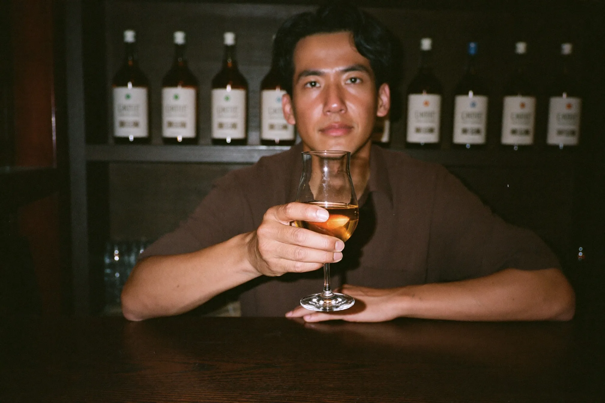 Man holding a plain unlabelled tulip glass of amber whisky at a dim timber bar counter in Kuala Lumpur