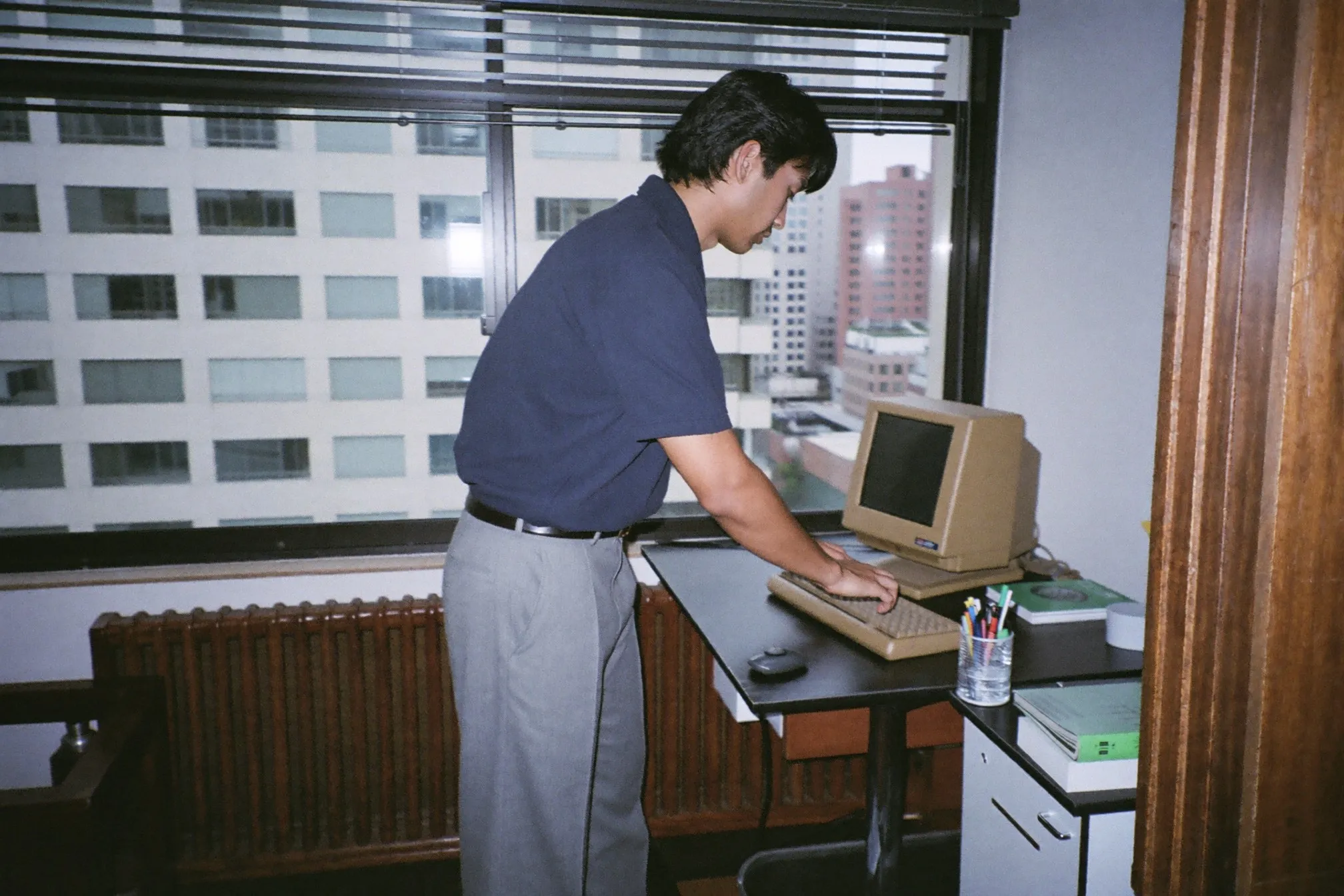 Man in a plain navy polo and soft grey trousers working at a desk by the window of a Kuala Lumpur condo